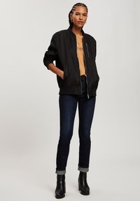 Czarna kurtka bomberka z żeberkowym kołnierzem, przednim zamkiem i bocznymi kieszeniami. Nosi się ją na beżowej koszuli, w zestawie z ciemnymi dżinsami i czarnymi kozaczkami na obcasie.