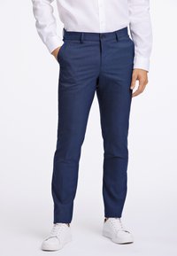 Navyblaue maßgeschneiderte Hosen mit einer subtilen Textur, ausgestattet mit seitlichen Taschen und einem Knopfverschluss, kombiniert mit weißen Sneakers.