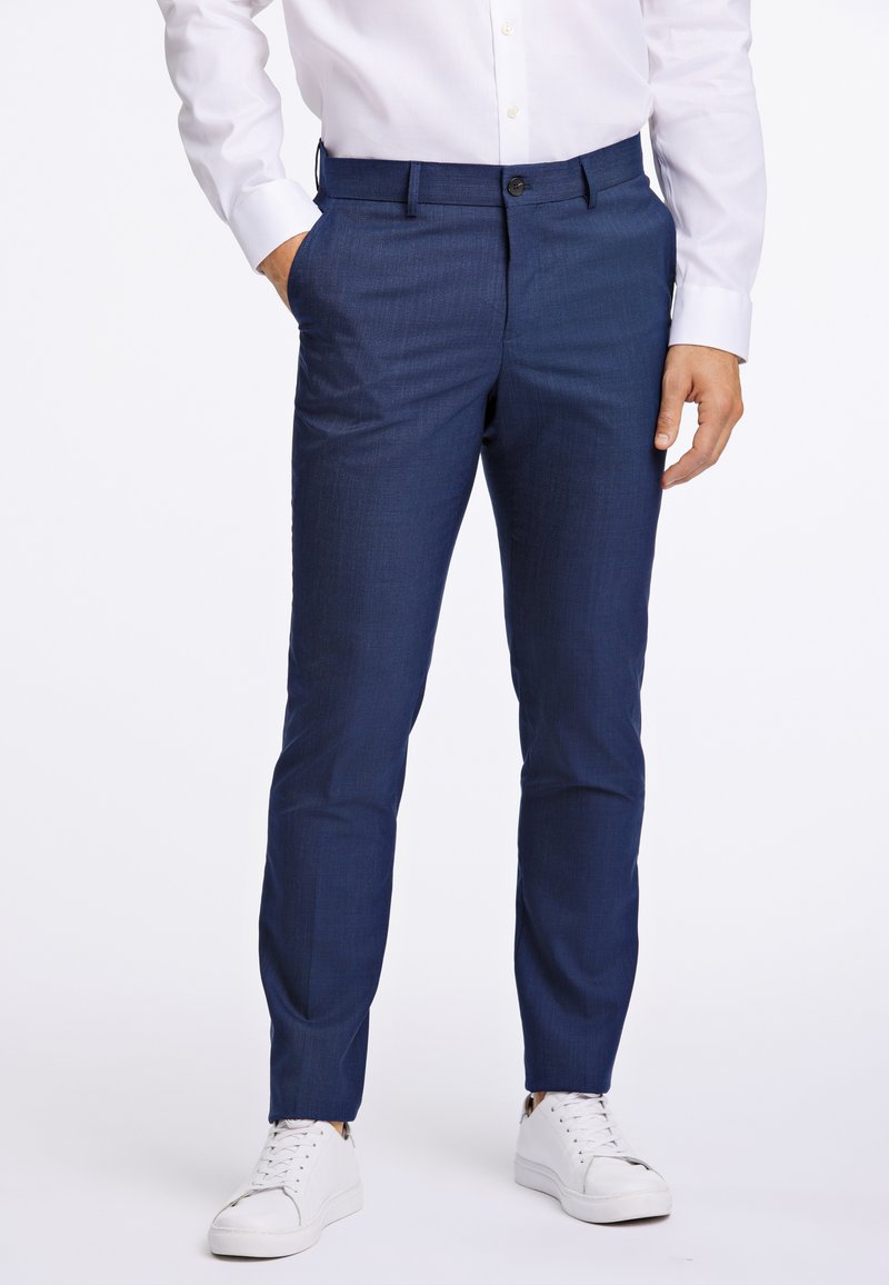 Navyblaue maßgeschneiderte Hosen mit einer subtilen Textur, ausgestattet mit seitlichen Taschen und einem Knopfverschluss, kombiniert mit weißen Sneakers.