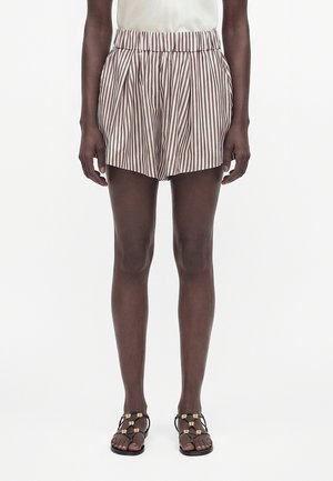 Osoba odjevena u bež i bijele vertikalno prugaste kratke hlače i crne sandale sa zlatnim detaljima, stoji ispred jednostavne svijetle pozadine.