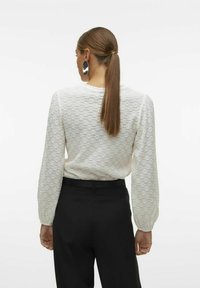 Top bianco texturizzato a maniche lunghe con motivo floreale, abbinato a pantaloni neri. I capelli sono raccolti in una coda di cavallo e sono visibili grandi orecchini d'argento.