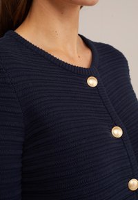 Navy ribbed cardigan with a round neckline, featuring golden button accents along the front and a textured knit pattern.