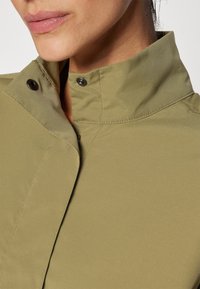 Olive zelená bunda s vysokým límcem, vybavená patentními knoflíky a hladkou, texturovanou látkou. Detaily zahrnují viditelné stehy na límci.