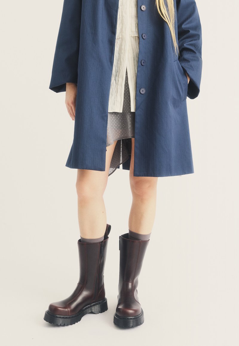 Une personne porte un manteau bleu marine par-dessus une robe blanche et une jupe grise transparente, associée à des bottes en cuir marron foncé mi-mollet et des chaussettes grises, debout sur un sol blanc.