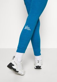 Teal, textured leggings with a white "LA SPORATIVA" logo on the calf, paired with white and black sneakers and white socks.