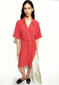 Rotes Wickelkleid mit kurzen Ärmeln, einem Kragen und einem Gürtel. Getragen mit schwarzen Schlippersandalen, während eine hellfarbige Jacke gehalten wird. Glatte Textur.