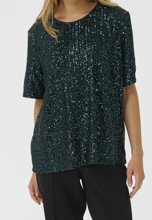 Femme portant un haut court à manches courtes bleu sarcelle foncé avec des sequins, assorti à un pantalon noir, debout, les bras détendus.