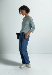 Lichtblauw top met driekwart mouwen, gecombineerd met losse blauwe jeans en witte sneakers, tegen een effen, lichtgekleurde achtergrond.