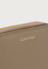 Gros plan d'un portefeuille taupe Calvin Klein avec une fermeture éclair dorée et le nom de la marque embossé sur une surface en cuir lisse.