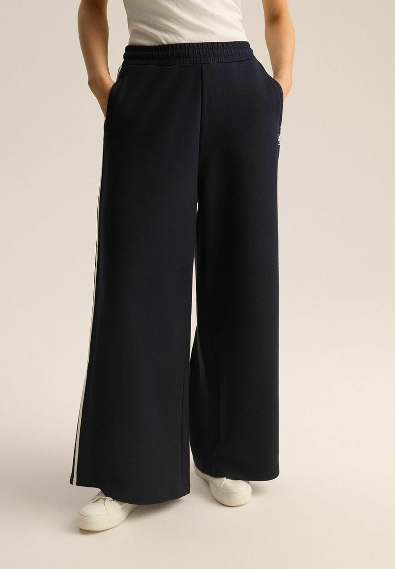 Granatowe dresy z szerokimi nogawkami, z elastycznym pasem, kieszeniami bocznymi i kontrastowymi paskami po bokach; zestawione z białymi sneakersami.