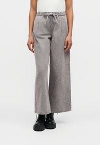 Άτομο που φοράει χαλαρό γκρι τζιν παντελόνι με ελαστική μέση και κορδόνι, συνδυασμένο με μαύρα πλατφόρμα μπότες σε λευκό φόντο.