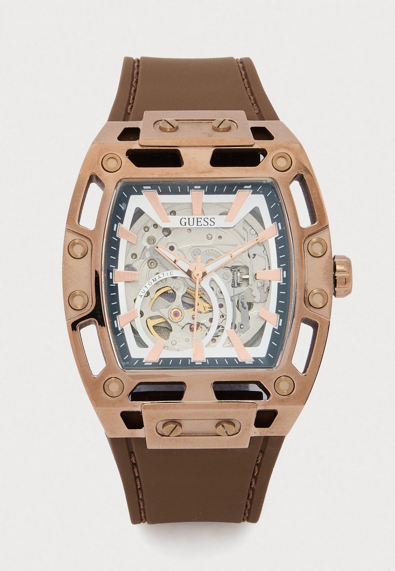 Orologio con cinturino in gomma marrone, cassa rettangolare tono bronzo, quadrante trasparente che rivela i meccanismi interni, dettagli rosa e marcatori delle ore audaci.