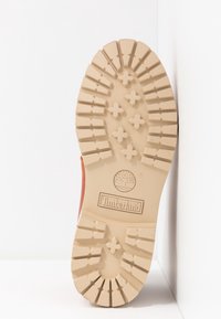 Semelle en caoutchouc beige avec des crampons profonds, des clous en forme de croix, et le logo Timberland embossé près du centre de la semelle.