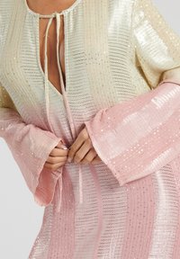 Glitterende jurk met een verloop van crème naar roze, met golvende texturen, lange klokmouwen en een striksluiting bij de hals.
