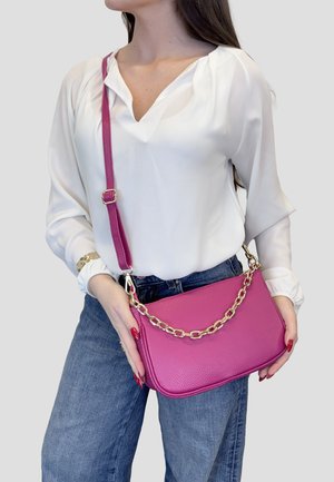 Vrouw met witte blouse en blauwe spijkerbroek houdt een roze leren handtas vast met een gouden ketting en verstelbare schouderriem.