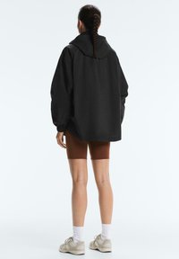 Person mit geflochtenem Haar, die eine lockere schwarze Jacke, braune Radhosen, weiße Socken und beige Turnschuhe trägt, steht mit dem Rücken zur Kamera vor weißem Hintergrund.
