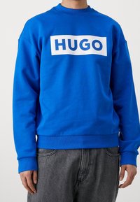 Sudadera azul hecha de una mezcla de algodón, con un logo rectangular blanco que lleva "HUGO" en letras negritas en el pecho. Puños y dobladillo acanalados.