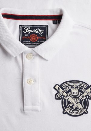 Biała koszulka polo wykonana z teksturowanej bawełny, z plisą na dwa guziki, haftowanym logo w ciemnoniebieskim kolorze oraz czerwonymi akcentami na kołnierzyku.