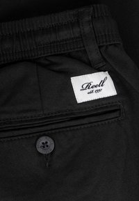 Primer plano del bolsillo trasero de pantalones negros con botón negro y etiqueta de tela blanca que dice "Reell est. 1997."
