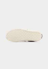 Semelle d'une basket noir et blanc avec une semelle extérieure en caoutchouc crème à motif gaufré, sur un fond blanc uni.