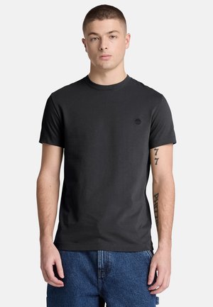 Giovane uomo che indossa una t-shirt nera semplice a maniche corte e jeans blu, in piedi con le braccia rilassate su uno sfondo neutro.