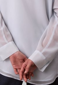 Blouse blanche avec manches longues transparentes et coupe droite. Les poignets sont en tissu uni, les mains sont jointes derrière le dos.