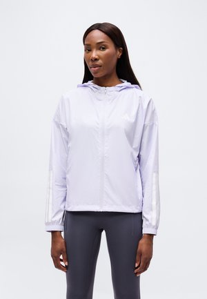 Donna in piedi con espressione neutra, indossa una giacca con cappuccio e zip di colore viola chiaro e leggings grigio scuro su uno sfondo semplice.