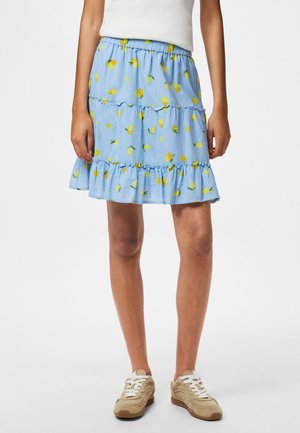 Lyseblåt knælængde skørt med gult citronprint, flæser i lag, båret med hvid top og beige sneakers på en stående person.