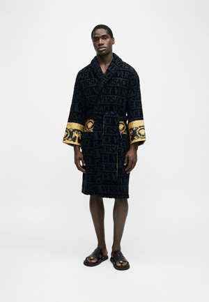 Homme portant une robe en velours noir avec le texte "VERSACE" embossé et des poignets brodés en or, associé à des sandales noires, debout sur un fond blanc.