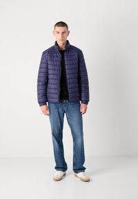 Marineblå quiltet jakke med høy krage, frontglidelås og elastiske ermer, kombinert med blå rette jeans og hvite joggesko.