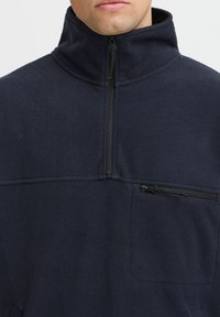 Pullover in pile di colore blu navy scuro, con colletto alto, chiusura a metà zip e tasca frontale con zip caratterizzata da una finitura testurizzata.