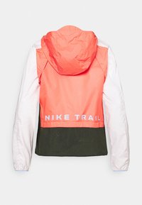 Nike Trail -takki, jossa korallinen yläosa, valkoiset hihat ja tummanvihreä alaosa. Takana oleva merkittävä valkoinen logoilu.