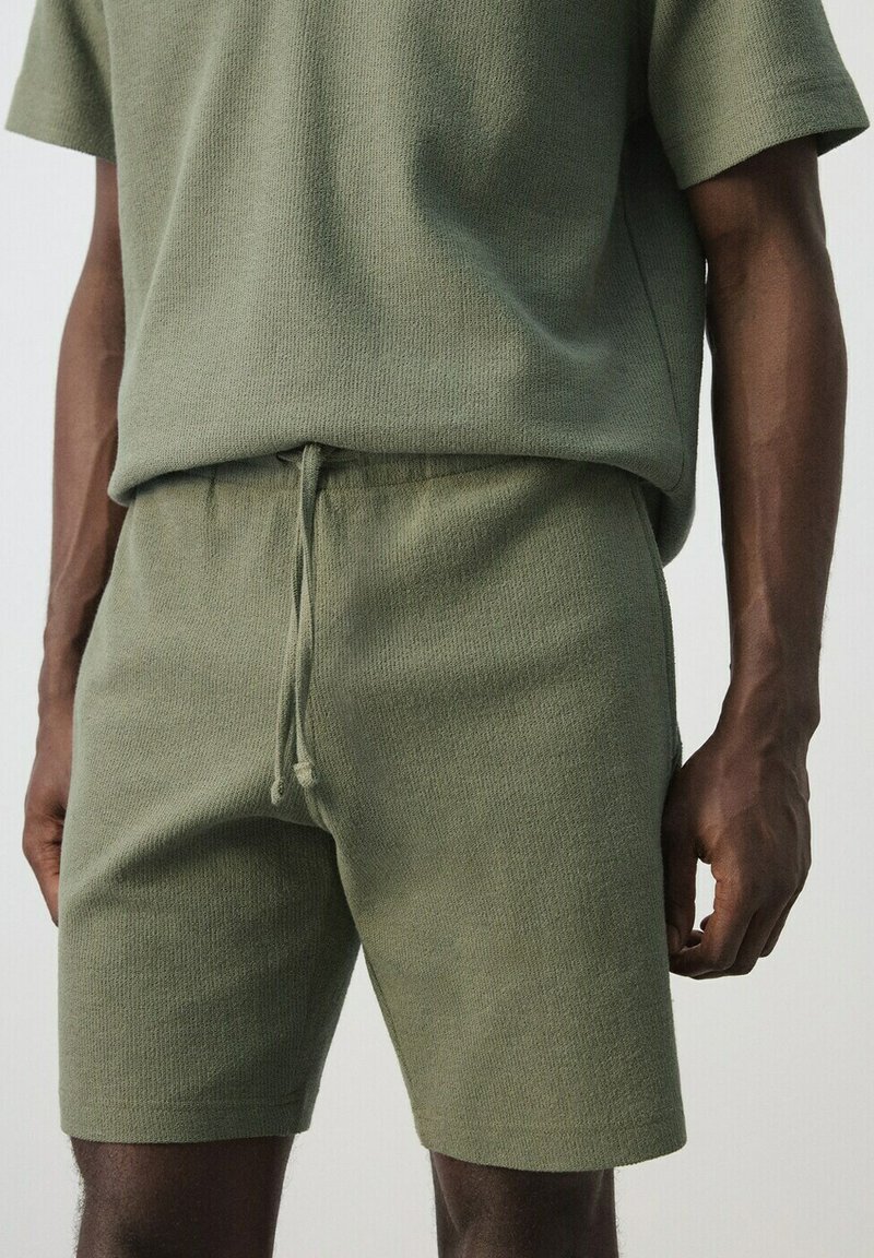 Homme portant un short en maille vert olive avec cordon de serrage et un haut assorti à manches courtes en maille vert olive, debout devant un fond uni.