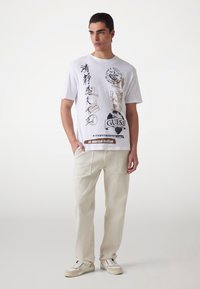 Vit t-shirt med olika svarta och guld grafiska tryck, kombinerad med ljusbeige byxor som har två framfickor och en avslappnad passform.