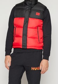 Homme portant une doudoune sans manches noire et rouge par-dessus une chemise noire à manches longues et un pantalon noir avec le texte "HUGO" en flammes sur la cuisse.