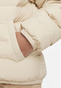 Chaqueta acolchada beige con un exterior liso y brillante, puños elásticos y un bolsillo lateral. La tela es suave y ligeramente acolchada.