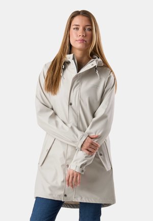 Mujer con cabello largo y liso que lleva un impermeable con capucha beige claro con botones de presión y jeans azules, de pie contra un fondo blanco.