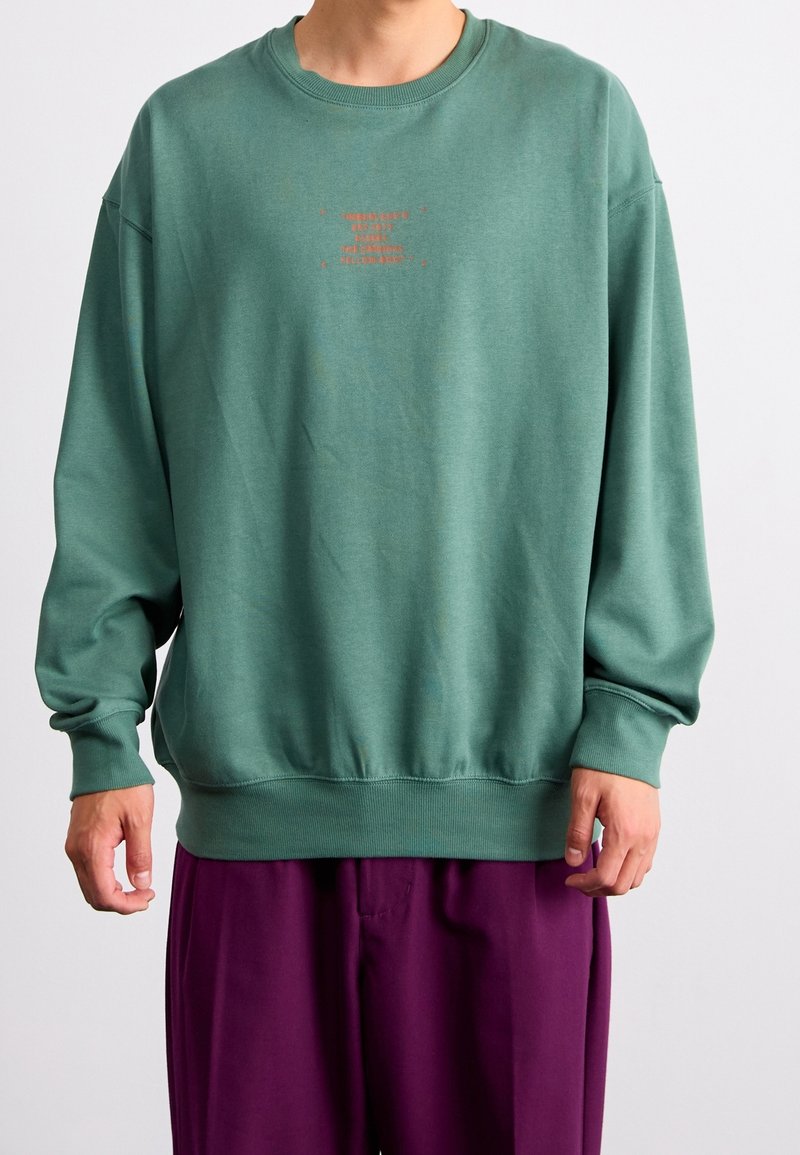Personne portant un sweat vert ample avec un petit texte orange sur la poitrine et un pantalon violet ample, debout devant un fond uni.