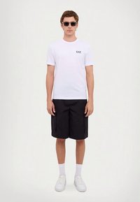 Mladý muž v černých slunečních brýlích, bílé tričko EA7, černé šortky s kapsami, bílé ponožky a bílé tenisky, stojící proti bílému pozadí.