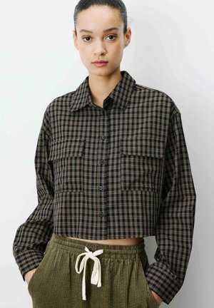 Femme portant une chemise courte à carreaux noirs et beiges à boutons avec manches longues et un pantalon en velours côtelé vert olive avec un cordon blanc.