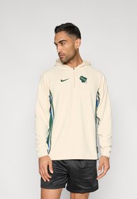 Béžová mikina s kapucňou a dlhými rukávmi, s zelenými a modrými bočnými akcentmi a logom "Bucks" na hrudi. Má zips na krku.