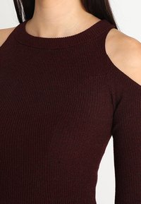 Top de punto acanalado burdeos que presenta un escote redondo y cortes en los hombros. La textura es suave, con mangas y cuerpo ajustados.