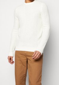 Witte gebreide trui met een textuurpatroon en ronde hals, gecombineerd met tan broek met ritsaccenten en zijzakken.