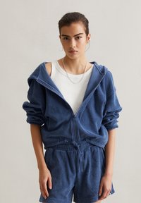 Felpa blu con zip e cappuccio realizzata in tessuto morbido, caratterizzata da maniche arrotolate e cordino nella parte inferiore, abbinata a pantaloni corti coordinati.