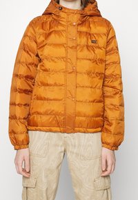 Veste matelassée orange à finition brillante, dotée d'une capuche et de boutons-pression, surface texturée et poignets élastiques.