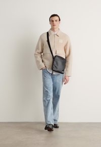 Chaqueta de forro polar beige con cremallera, detalles de bolsillo, jeans azul claro y un bolso bandolera negro. Combinada con zapatos negros, de diseño minimalista.