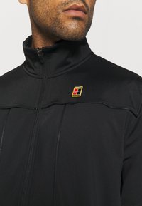 Chaqueta deportiva negra con cuello alto, cierre de cremallera y un pequeño logo colorido en el hombro. Cuenta con una tela suave y elástica.