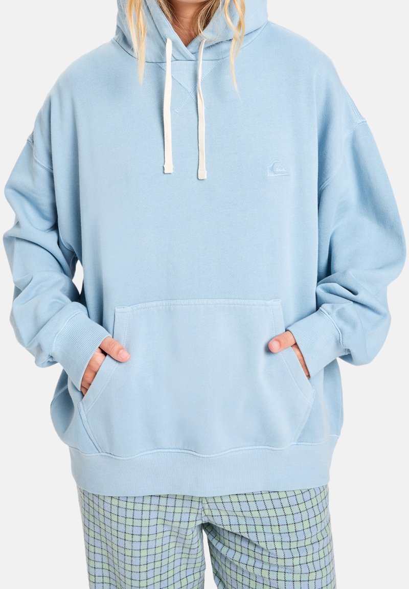 Personne portant un sweat à capuche bleu clair oversize avec des cordons blancs et un pantalon vert à carreaux, les mains dans la poche avant du sweat.