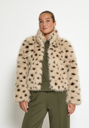 Kvinde iført en beige faux fur-jakke med sorte pletter over et olivengrønt outfit, stående mod en ensfarvet hvid baggrund.