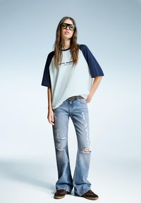Světle modré oversized tričko s potiskem loga a námořnickými raglánovými rukávy, doplněné o roztrhané světle modré zvonové džíny a hnědé boty.